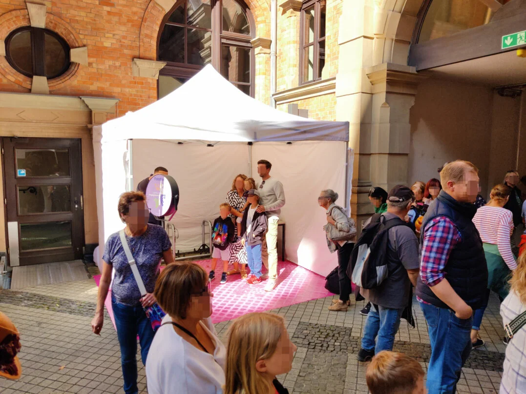 Eine Familie posiert für ein Foto in einer Fotobox, die unter einem weißen Zelt auf dem Stadtfest Wiesbaden am Rathaus aufgestellt ist. Im Vordergrund warten weitere Besucher in einer Schlange, um ebenfalls ein Foto zu machen.
