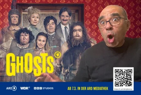 Ein Mann mit Brille macht ein überraschtes Gesicht, während er für ein Fotobox-Foto mit dem "Ghosts"-Plakat der ARD posiert. Das Plakat zeigt die Charaktere der Serie im Hintergrund.