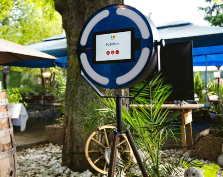 Eine akku-betriebene Fotobox steht draußen unter einem Baum in einem Garten, der für eine Veranstaltung dekoriert wurde. Im Hintergrund sind Sonnenschirme und Tische zu sehen.