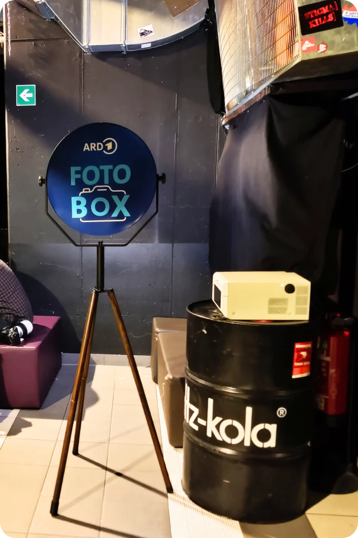 Ein Mann sitzt neben einer Fotobox, die für eine Party in einem Club aufgestellt wurde. Im Hintergrund sind Bildschirme zu sehen, während eine Wi-Fi-Verbindung den kabellosen Druck ermöglicht.