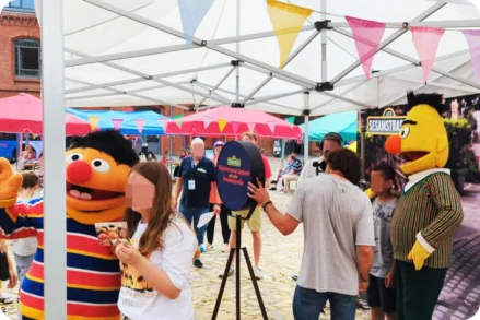 Besucher, darunter Kinder und Jugendliche, posieren lachend an einer Fotobox auf einem Familienfest. Die Figuren Ernie und Bert aus der Sesamstraße sind ebenfalls anwesend. Im Hintergrund sind weitere Besucher und Stände unter Zelten zu sehen.