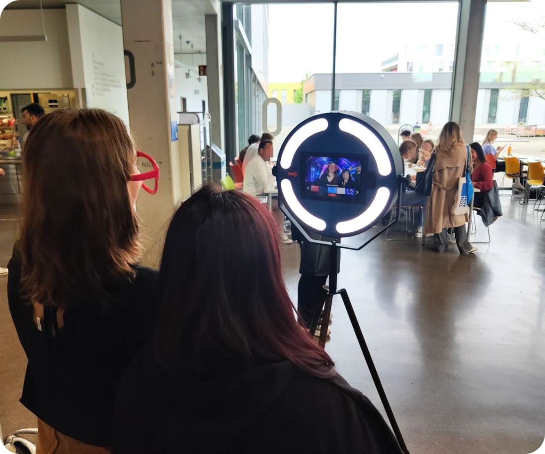 Zwei Besucherinnen posieren für ein Foto an einer Fotobox auf einem Event im Inneren der Hochschule Mainz. Im Hintergrund sind weitere Besucher und Tische in einem großen, hellen Raum zu sehen.