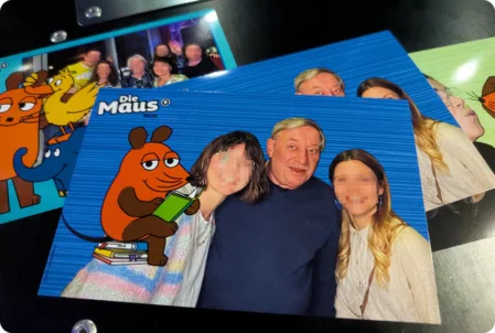 Zwei gedruckte Fotos von einem "Die Maus"-Event liegen auf einer Oberfläche. Die Bilder zeigen Gäste, die vor einem blauen Hintergrund mit den Figuren der Sendung "Die Maus" posieren.
