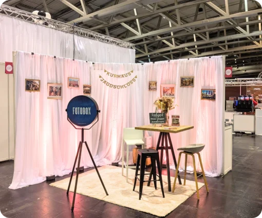 Ein ansprechender, kleiner Messestand mit einer Fotobox und einer Theke mit zwei Stühlen ist zu sehen. Im Hintergrund hängen gerahmte Bilder an einer weißen Wand.