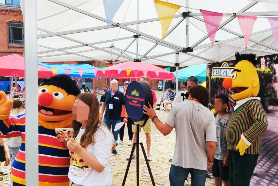 Besucher, darunter Kinder und Jugendliche, posieren lachend an einer Fotobox auf einem Familienfest. Die Figuren Ernie und Bert aus der Sesamstraße sind ebenfalls anwesend. Im Hintergrund sind weitere Besucher und Stände unter Zelten zu sehen.
